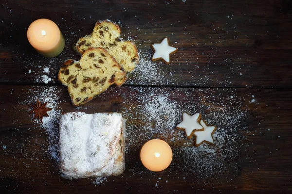 Christmas cake, traditional german criststollen sprinkled with powdered sugar, burning white candles and cinnamon star cookies on a dark rustic wooden board, top view from above, copy space — Stock Photo, Image