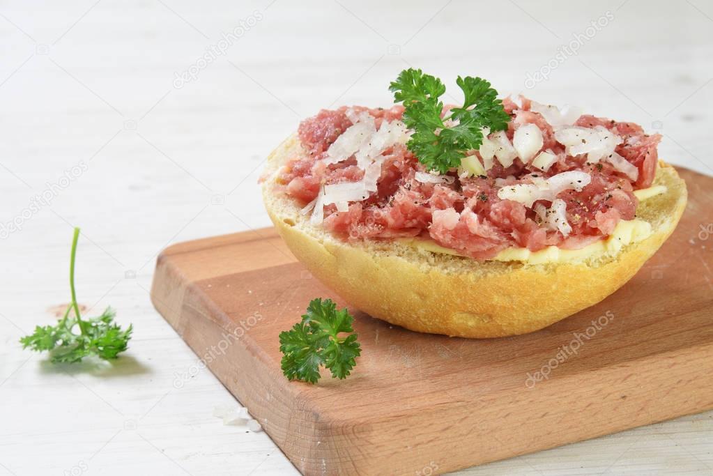 Bun with minced pork sausage, in germany called zwiebelmett with onions and parsley garnish on a breakfast board,  white wooden background with copy space, close up