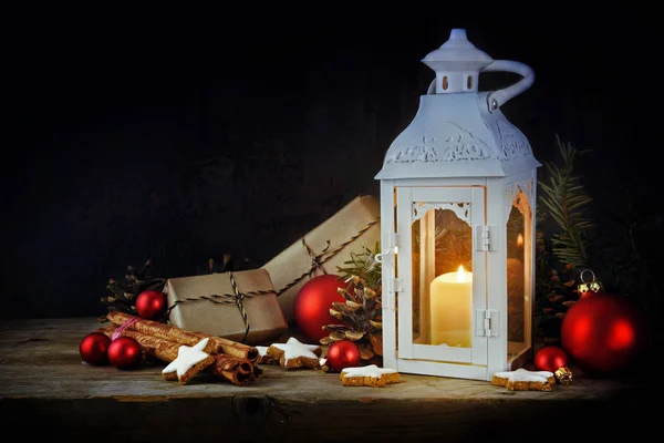 Karácsonyi dekoráció lámpa és gyertyafényes, ajándékok, a piros golyó, a fenyő kúpok és a fahéj csillag a cookie-k egy rusztikus, fából készült asztal, sötét háttér-val másol hely — Stock Fotó