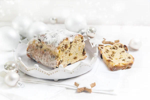 Deutscher Christstollen, genannt Cristollen mit Rosinen und kandierten Früchten auf weißem Teller, verziert mit hellen Weihnachtskugeln und Zimtsternen auf weißem Tisch, Kopierraum — Stockfoto