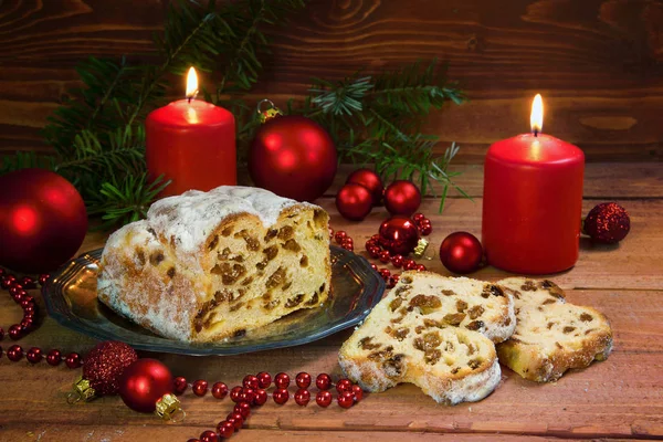 Festive christmas cake, german criststollen with raisins and almonds, decorated with burning candles, red christmas balls and fir branches on a rustic wooden background — Stock Photo, Image