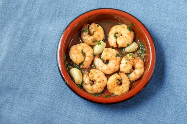 Shrimps or prawns and garlic in olive oil with parsley in a brown tapas bowl, spanish appetizer on a blue tablecloth background, flat top view from above, copy space — Stock Photo, Image
