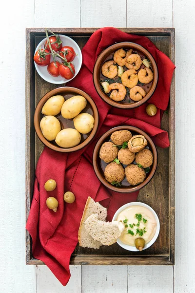 Spanish tapas finger food, baked olives, prawn shrimps, potatoes, tomato and garlic dip served as party appetizers on a dark wooden tray with a red napkin on white painted wood, flat top view from above — Stock Photo, Image