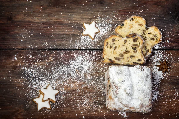 Weihnachtlicher Hintergrund, traditioneller deutscher Christstollen mit Puderzucker, Zimtplätzchen und Anissternen bestreut auf dunklem, rustikalem Holzbrett, Draufsicht von oben — Stockfoto