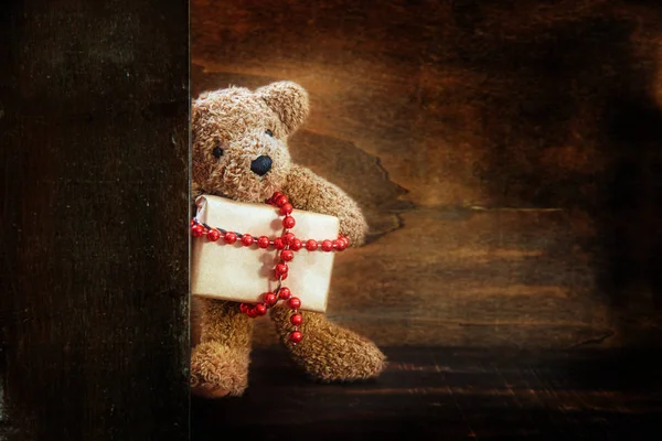 Mignon petit ours en peluche demi derrière un mur en bois apporte un cadeau de Noël décoré d'une chaîne à billes rouges, fond en bois rustique foncé avec espace de copie — Photo