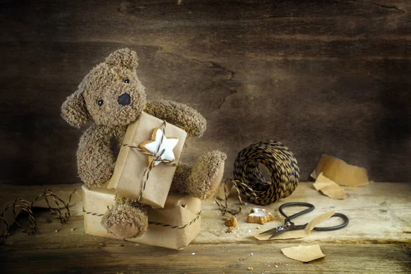 Ours en peluche assis sur une boîte-cadeau et montre son cadeau de Noël enveloppé, papier kraft, ruban et ciseaux, fond en bois rustique foncé avec espace de copie — Photo