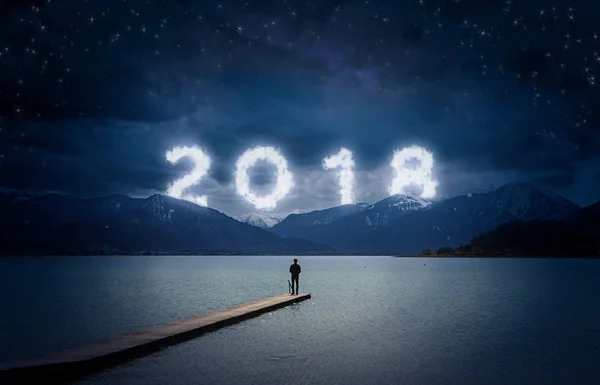 Fundo do ano novo, jovem de pé em um molhe em um lago e olhando para as montanhas sob o céu escuro com texto nublado 2018 — Fotografia de Stock