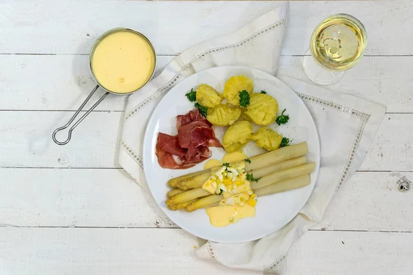 Asperge schotel met aardappelen, ham, saus en witte wijn van bovenaf op wit geschilderd hout — Stockfoto