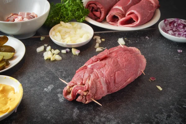 Preparação de rouladas de carne bovina, carne crua fresca enrolada recheada com mostarda, cebola, pepino em conserva e bacon em uma bancada de cozinha de ardósia escura — Fotografia de Stock