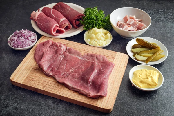 Preparación de paseos de ternera con los ingredientes de mostaza, cebolla, pepinos en escabeche, tocino y carne fresca en una tabla de cortar de madera y una encimera de cocina oscura —  Fotos de Stock