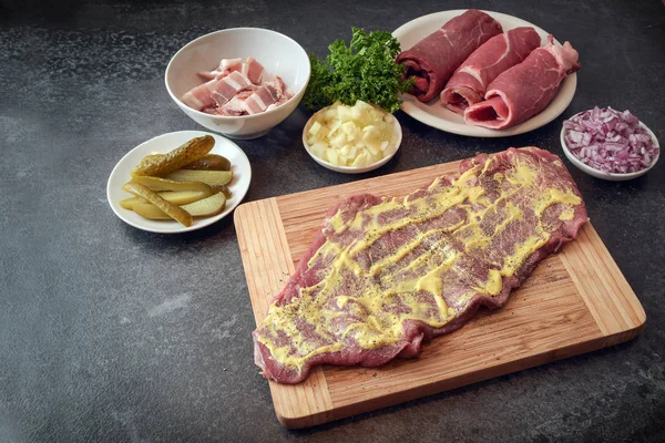 Preparar roulades de carne de res, carne cruda recubierta de mostaza e ingredientes como cebollas, pepino en escabeche y tocino en una tabla de cortar de madera, encimera de cocina oscura con espacio para copiar — Foto de Stock