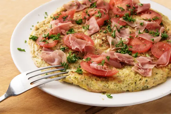 Omelette aux tomates, jambon de prosciutto et garniture de persil et une fourchette sur une assiette blanche sur une table en bois rustique, fermer — Photo