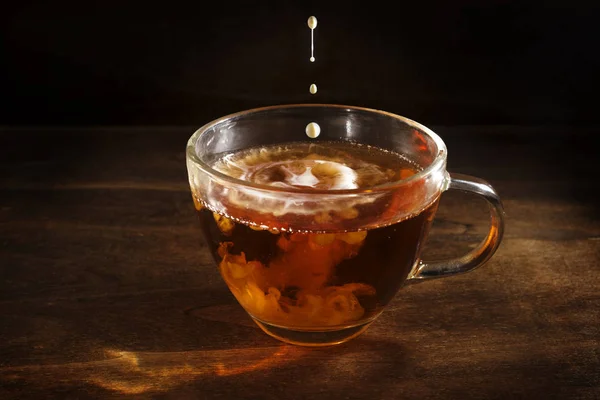 Tè nero appena preparato in una tazza di vetro e alcune gocce di latte o crema su uno sfondo di legno rustico scuro, bevanda calda contro il freddo e l'influenza, spazio copia — Foto Stock