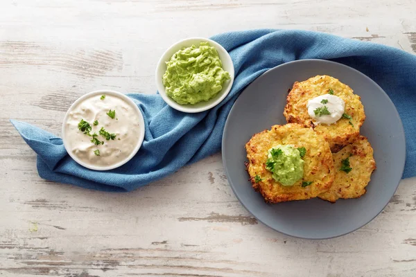 Rosti di verdure fritte di cavolfiore e parmigiano con due tuffi di panna acida e avocado, guarnizione di prezzemolo, piatto blu e tovagliolo su un tavolo di legno luminoso con spazio copia, vista ad angolo alto dall'alto — Foto Stock