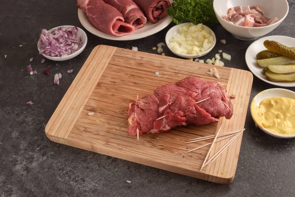 Preparação de rouladas de carne bovina, carne crua fresca recheada com mostarda, cebola, pepino em conserva e bacon em uma placa de corte de madeira, bancada de cozinha de ardósia escura — Fotografia de Stock