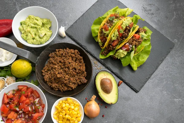 Tacos messicani ripieni e ingredienti come carne macinata fritta, salsa di pomodoro, guacamole, mais e spezie su un piatto di ardesia scura, vista dall'alto — Foto Stock