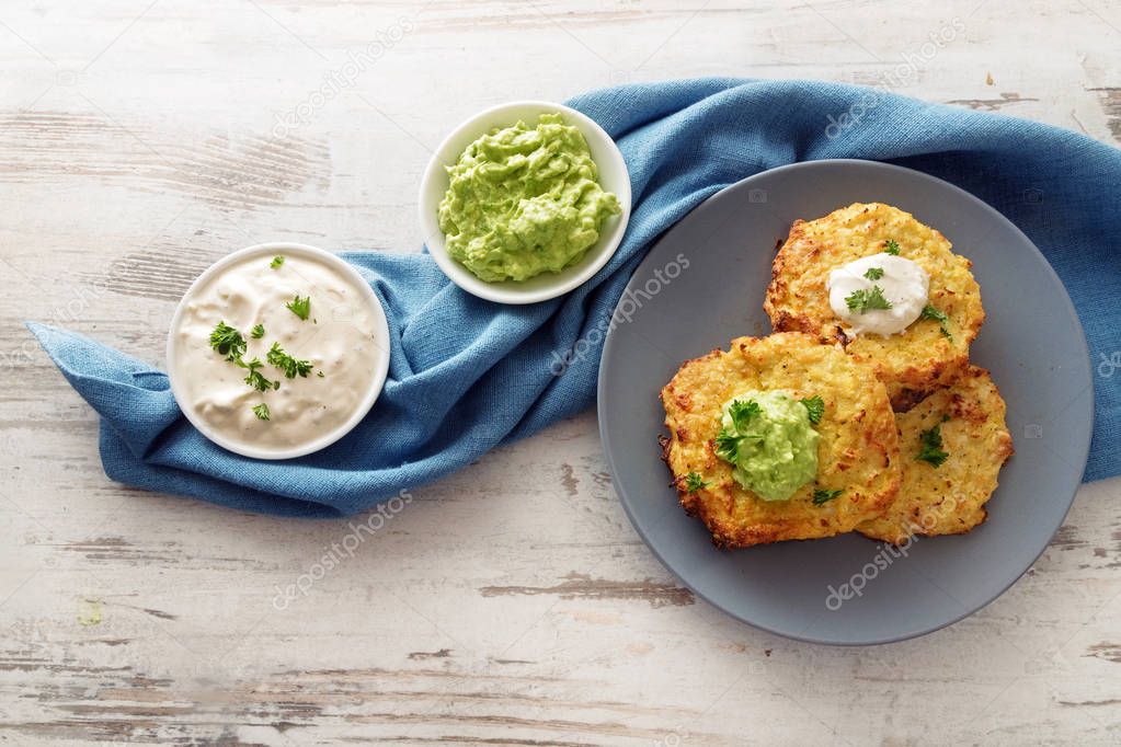 fried vegetable rosti from cauliflower and parmesan cheese with two dips from sour cream and avocado, parsley garnish, blue plate and napkin on a bright wooden table with copy space, high angle view from above