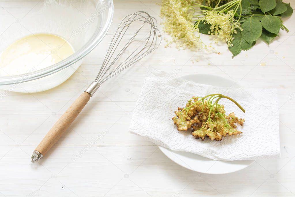 making fried elderflower pancakes, whisk, bowl with dough and finished pastry on a white wood background
