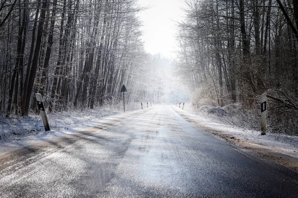 Ijzige landweg leidt door een bos van de winter met kale bomen en sneeuw, concept voor veiligheid, verkeer en vervoer in het koude seizoen — Stockfoto