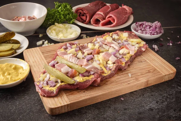 Preparing beef roulades on a wooden cutting board, fresh raw meat with mustard, onions, pickled cucumber and bacon, dark kitchen countertop — Stock Photo, Image