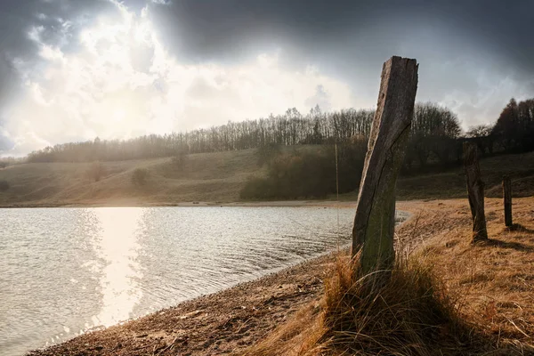 Alte Holzpfähle am Ufer eines Sees mit Sonnenreflexen, Landschaft im Abendlicht unter dunklen Wolken, Kopierraum, Fahrt an der Ostsee bei Lübeck — Stockfoto