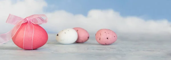 Uova di Pasqua rosa e bianche, una con un nastro contro un cielo blu sfocato con nuvole in formato banner panoramico, copiare lo spazio — Foto Stock