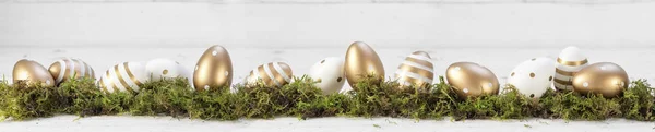 Easter decoration, gold painted eggs on moss against a gray white wooden background, extra wide panoramic banner, copy space — Stock Photo, Image