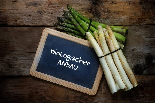 Зеленая и белая спаржа и доска с немецким текстом biologischer anbau, что означает органическое земледелие, на деревенском деревянном фоне, вид сверху, пространство для копирования — стоковое фото