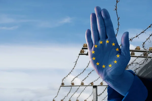 Mano con bandiera europea ferma l'immigrazione dei rifugiati, recinzione di confine sfocata sullo sfondo, cielo blu con spazio copia — Foto Stock