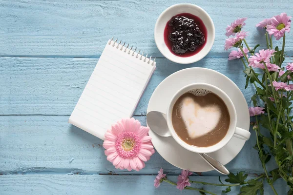 Taza de café con un corazón de espuma de leche, flores de color rosa y una almohadilla de escritura sobre un fondo de madera azul pastel con espacio para copiar —  Fotos de Stock