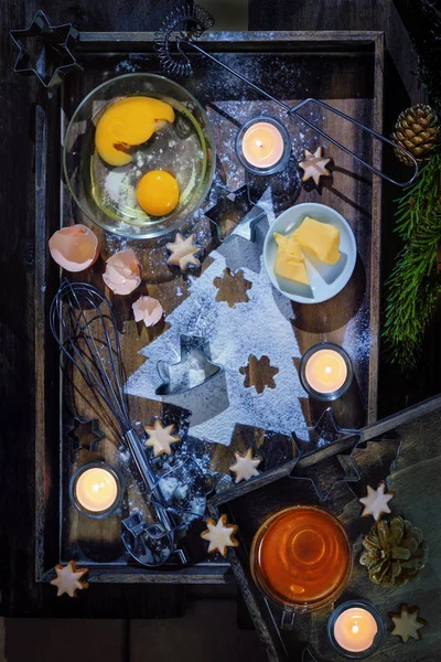 Cuocere biscotti di Natale, ingredienti, una forma di abete da farina, utensili e candele accese su un vassoio di legno scuro, posa piatta, vista ad alto angolo dall'alto — Foto Stock