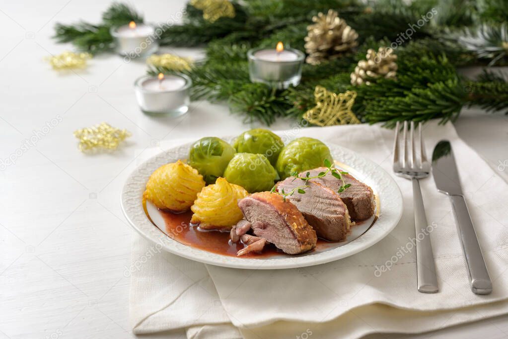 Seared duck breast with brussels sprout, duchess potatoes and sauce as a festive dinner, served on a white wooden table with candles fir branches and Christmas decoration