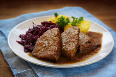 Soslu haşlanmış biftek, patates ve kırmızı lahana, beyaz tabakta servis edilir ve ahşap bir masada mavi peçete.