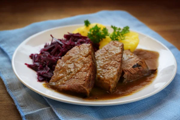 Duszona wołowina z sosem, ziemniakami i czerwoną kapustą, podawana na białym talerzu i niebieska serwetka na rustykalnym drewnianym stole — Zdjęcie stockowe