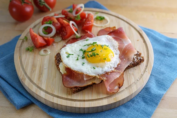 Ekmek, domates, soğan halkaları, garnitür, mutfak tahtası ve mavi peçeteyle servis edilen sahanda yumurta ve jambon. — Stok fotoğraf