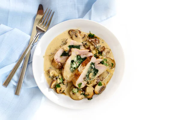 Gefülltes Hühnerbrustfilet mit Spinat, Pilzen und einer cremigen Gorgonzola-Sauce auf einem Teller, blaue Serviette und Besteck auf weißem Hintergrund mit Kopierraum, Blick von oben — Stockfoto