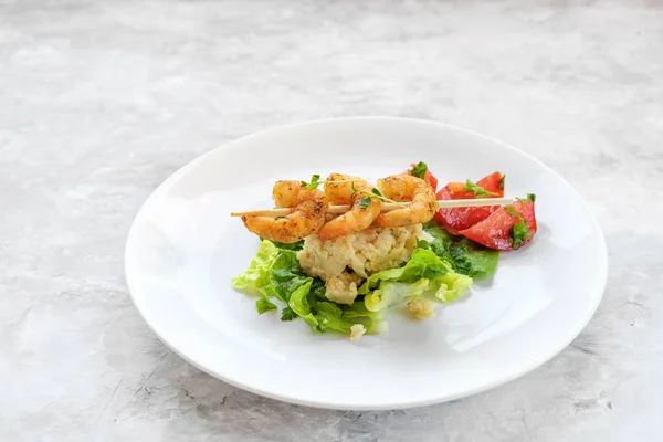 Шашлыки с креветками на ризотто из цветной капусты, салат и помидоры на белой тарелке, светло-серый камень с копировальным пространством — стоковое фото