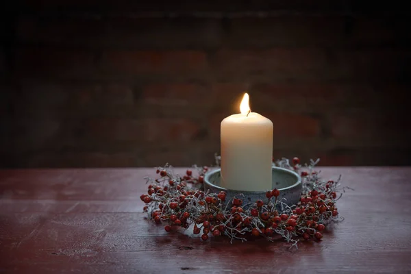 Kırmızı ahşap bir masada, gül kalçalarındaki küçük bir çelenkle yanan mum ve gümüş yastık çalısı, Advent, Noel ve Yeni Yıl için kış süsü, fotokopi alanı olan karanlık bir arka plan. — Stok fotoğraf