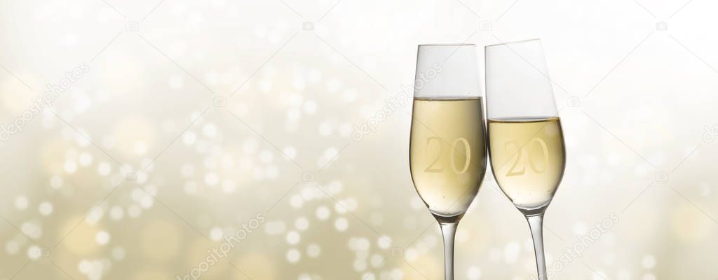 Happy New Year 2020, two champagne glass flutes toasting against a light golden bubble bokeh background, party concept in panoramic format with copy space