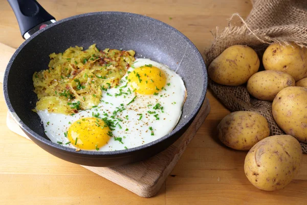 Fried eggs and Swiss roesti in a pan on a wooden kitchen table with a sack of potatoes — 스톡 사진