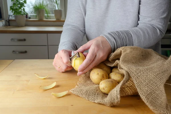 As mãos de uma mulher estão descascando batatas cruas em uma mesa de madeira na cozinha em casa — Fotografia de Stock