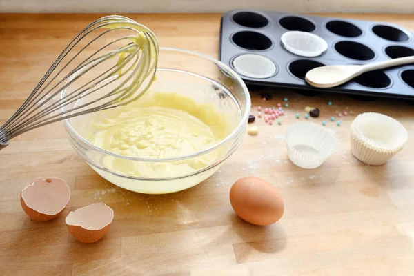 Whisk com massa de cupcake, cascas de ovos e uma lata de muffin em uma placa de cozinha de madeira, conceito de cozimento — Fotografia de Stock