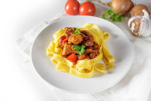 Gulasch eller gryta från fläsk med lök och paprika på tagliatelle pasta serveras på en tallrik och ingredienser på ett vitt bord, kopiera utrymme — Stockfoto