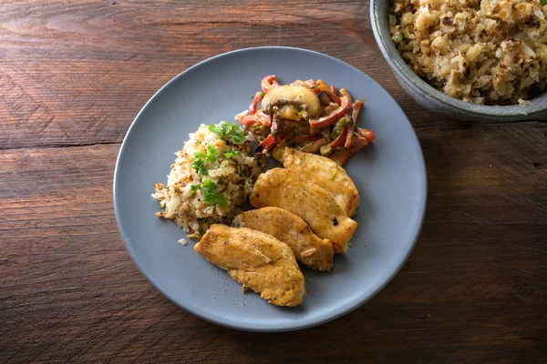 Chicken filet with cauliflower rice and vegetable ragout from bell pepper and mushroom, healthy diet meal on a blue plate on a dark rustic wooden table from above, copy space — Stock Photo, Image