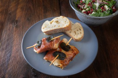 Kızarmış tavuk saltimbocca jambonlu ve adaçayı yapraklı koyu sade ahşap bir masada ekmek ve salata ile servis edilir.