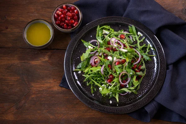 Ensalada de rúcula o cohete con granada, cebolla roja y parmesano servido en un plato oscuro, tazones pequeños con semillas y aderezo, fondo rústico de madera, espacio para copiar — Foto de Stock