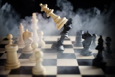 Satranç kraliçesi satranç tahtasındaki diğer taşların arasında kralı yendi, savaş boyunca çok duman, karanlık bir arka plan, saldırganlık konsepti, başarı ve takımlar arasındaki rekabet