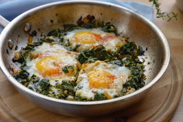 Кухонна сковорода зі шпинатом та смаженими яйцями, спеціями та трав'яним гарніром, здорова страва для низьковуглецевого раціону на дерев'яній дошці, крупним планом — стокове фото