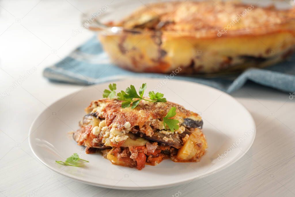 Moussaka, traditional Greek baked casserole of eggplants, potatoes, minced meat and tomatoes served with parsley garnish on a plate on a white table, selected focus, narrow depth of field