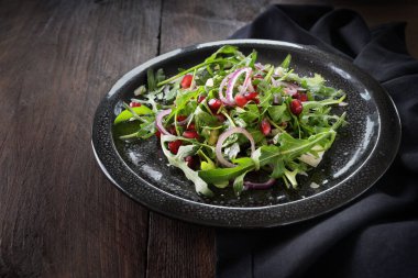 Taze roket salatası ya da roka, nar tohumu, kırmızı soğan ve beyaz peynir, siyah benekli bir tabakta, ahşap bir masada, kopyalama alanı, seçilmiş odak, dar bir alan derinliği.